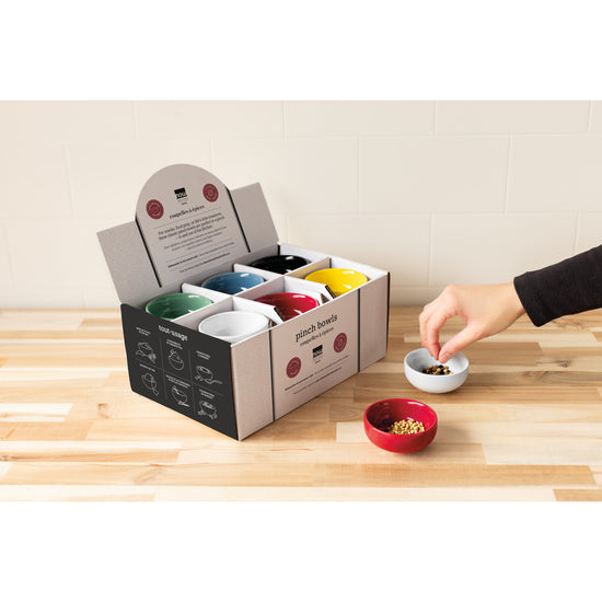 A hand is reaching into a white pinch bowl next to a counter display unit of different colored pinch bowl by Now Designs.